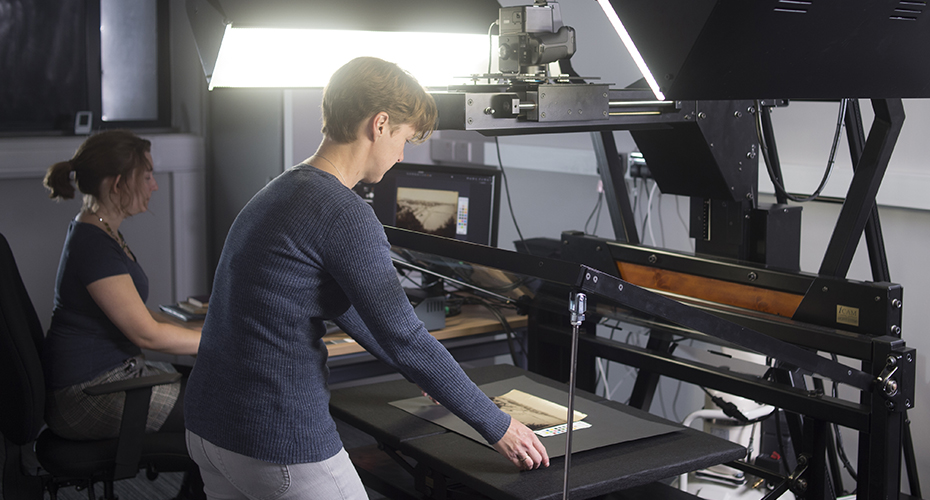 An image of two people using the copystand in Lab 1 to digitise flat material - one adjusts the material to make sure it is in the right place, and one uses the computer to check quality of the image once it is taken. 