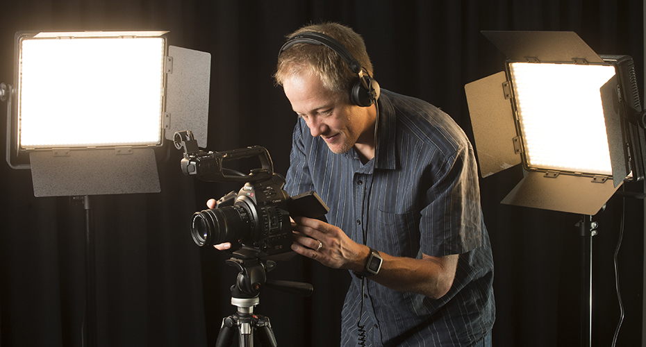 An image of someone in a studio with headphones on and lighting behind them filming using a videocamera. 
