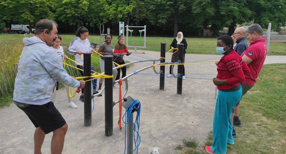 A group exercise session in a park. Participants are pulling on elastic straps attached to a piece of outdoor gym equipment.