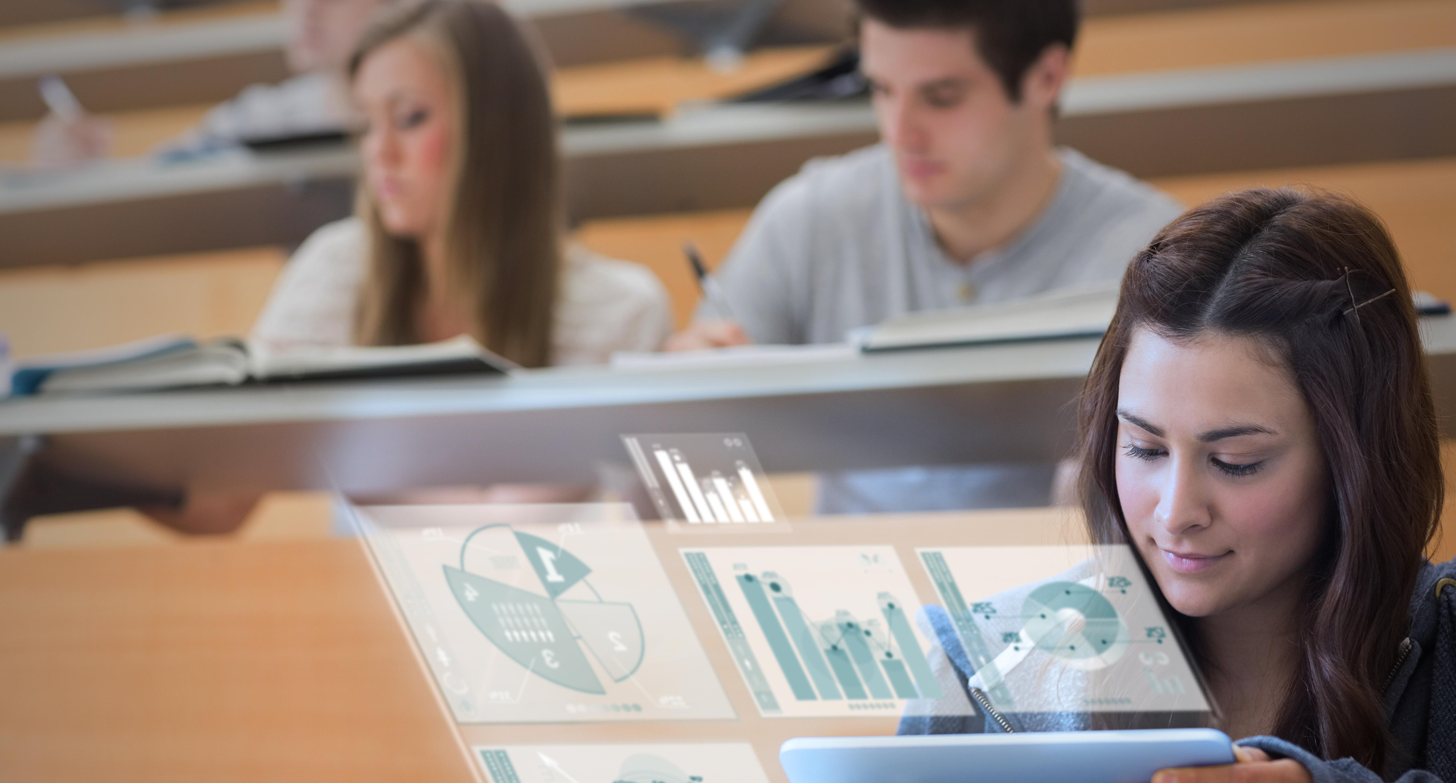 Student in a lecture theatre looking at a laptop with two other students in the row of seating behind, who are are writing notes. Superimposed on the image are data charts and graphs.