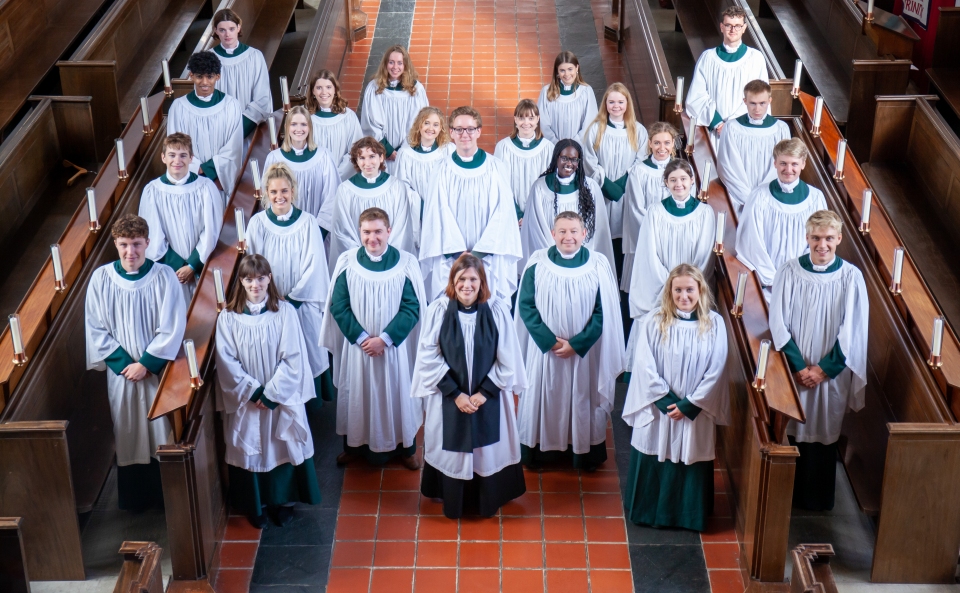 24 choristers of the Chapel Choir for the 2022-23 academic year stand smiling up towards the camera in the organ loft in the Mary Harris Chapel. They are accompanied by the Chaplain, the Rev'd Hannah Alderson; the Director of Chapel Music, Mr Michael Graham; and the Chapel Organist, Mr David Davies.