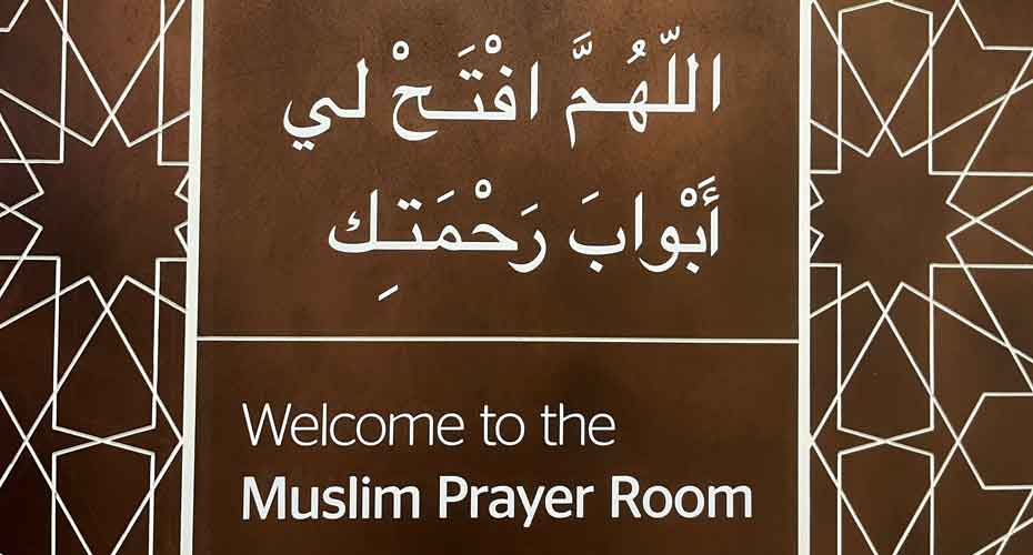 Brown sign with white writing in arabic and english reading Welcome to the muslim prayer room