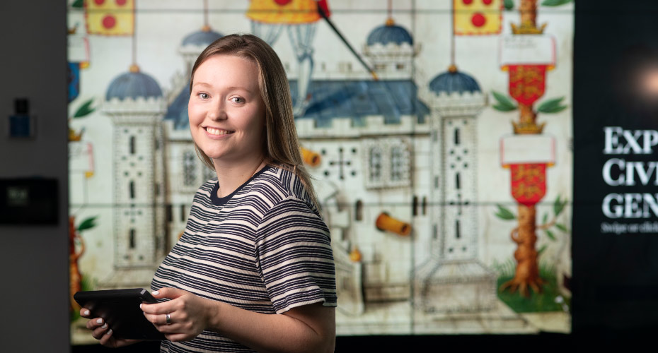 Girl smiling and standing with a tablet, in front of a large presentation screen showing an image of a historic castle