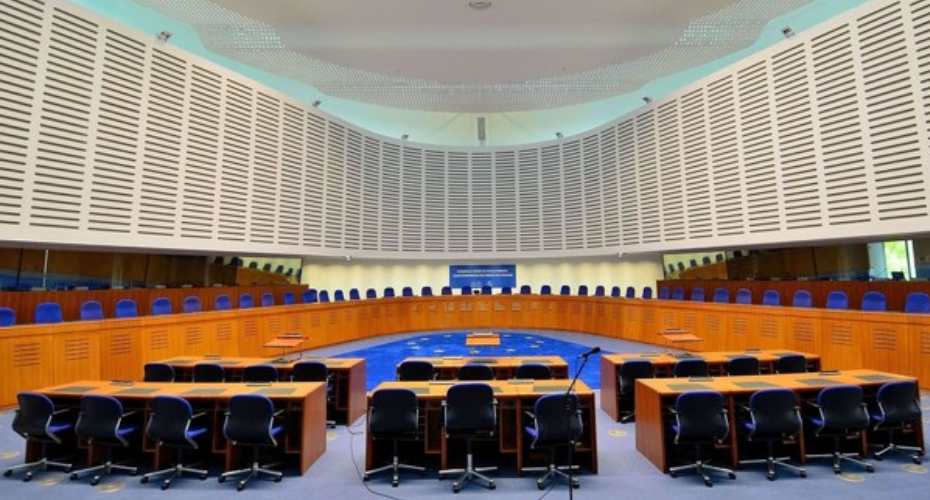 the inside of the European Court of Human Rights. There is an arc of brown desks with blue seats behind them. The tall white wall behind is slatted.
