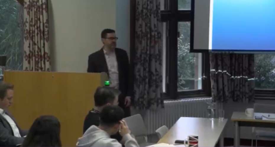A person stood in front of a bay window giving a lecture in a room with a large screen and some students sat at a table.