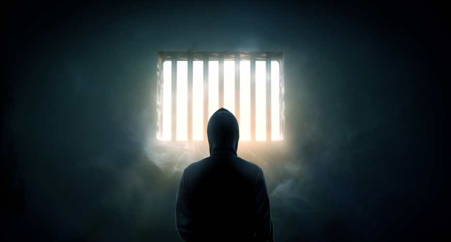 A silhouetted figure stands in front of a barred window through which light shines