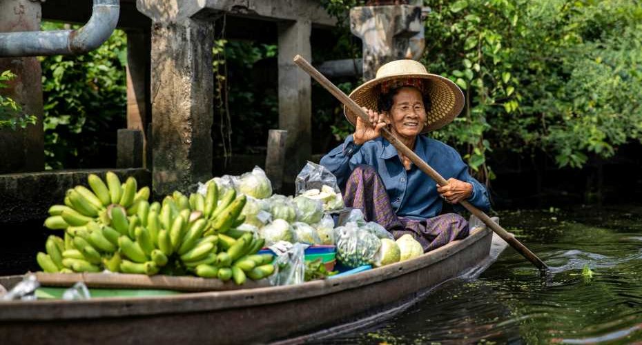 A woman sat in a small open vessel with a wooden oar in her hands. SHe has a large bunches of bananas in the boat.