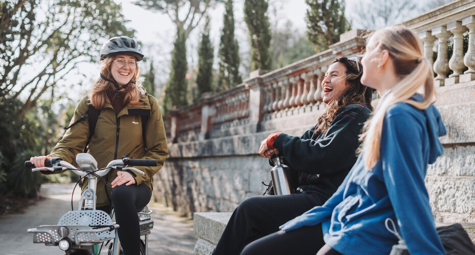 Three students laughing together. One has a bicycle with her and is wearing a helmet.