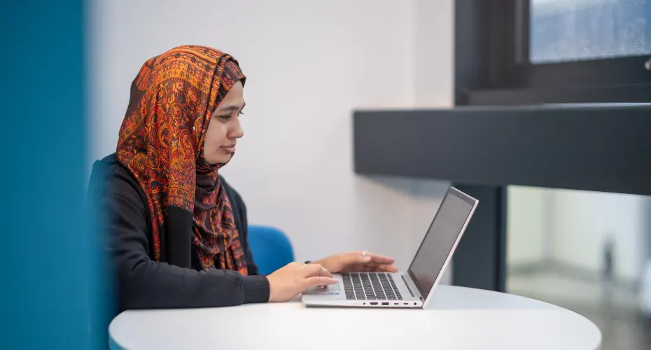 PhD student sitting at a desk with laptop open, in a study space in Penryn Campus.