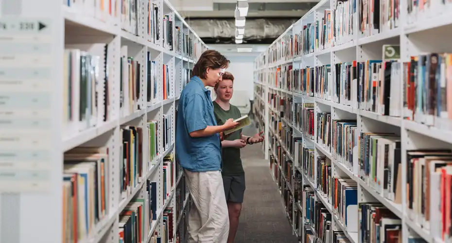 2 students standing looking at books in between two shelves of books in a library.