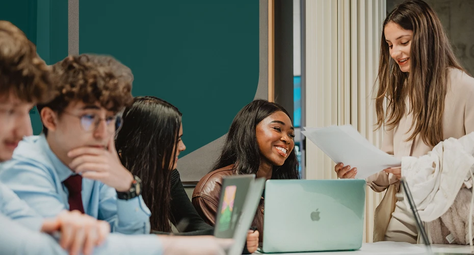 Students with laptops smiling and talking, student holding a piece of paper on the right