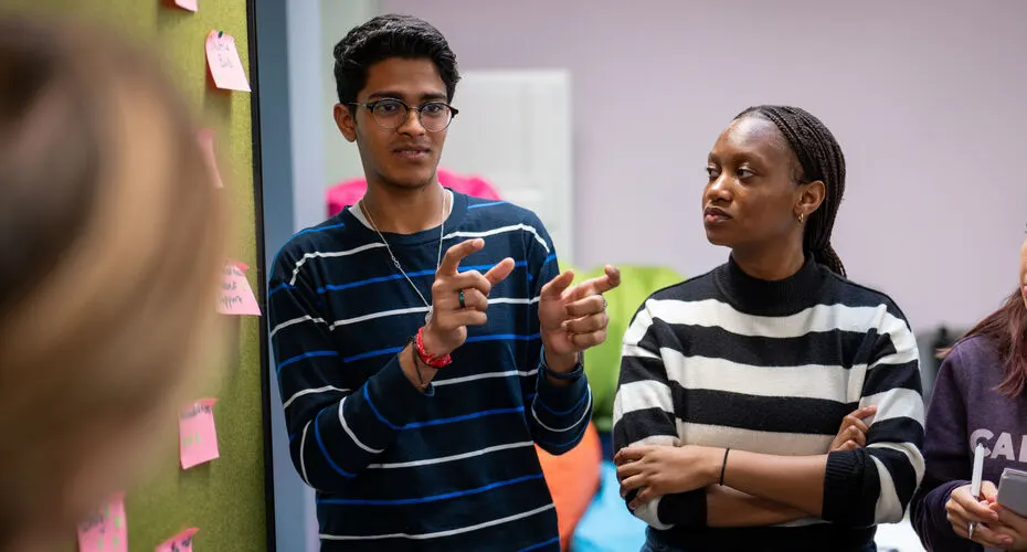 Two students standing together with one talking to a group and the other looking at them listening.