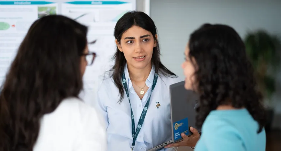 A student converses with two others in front of a colorful poster, engaged in a lively discussion.