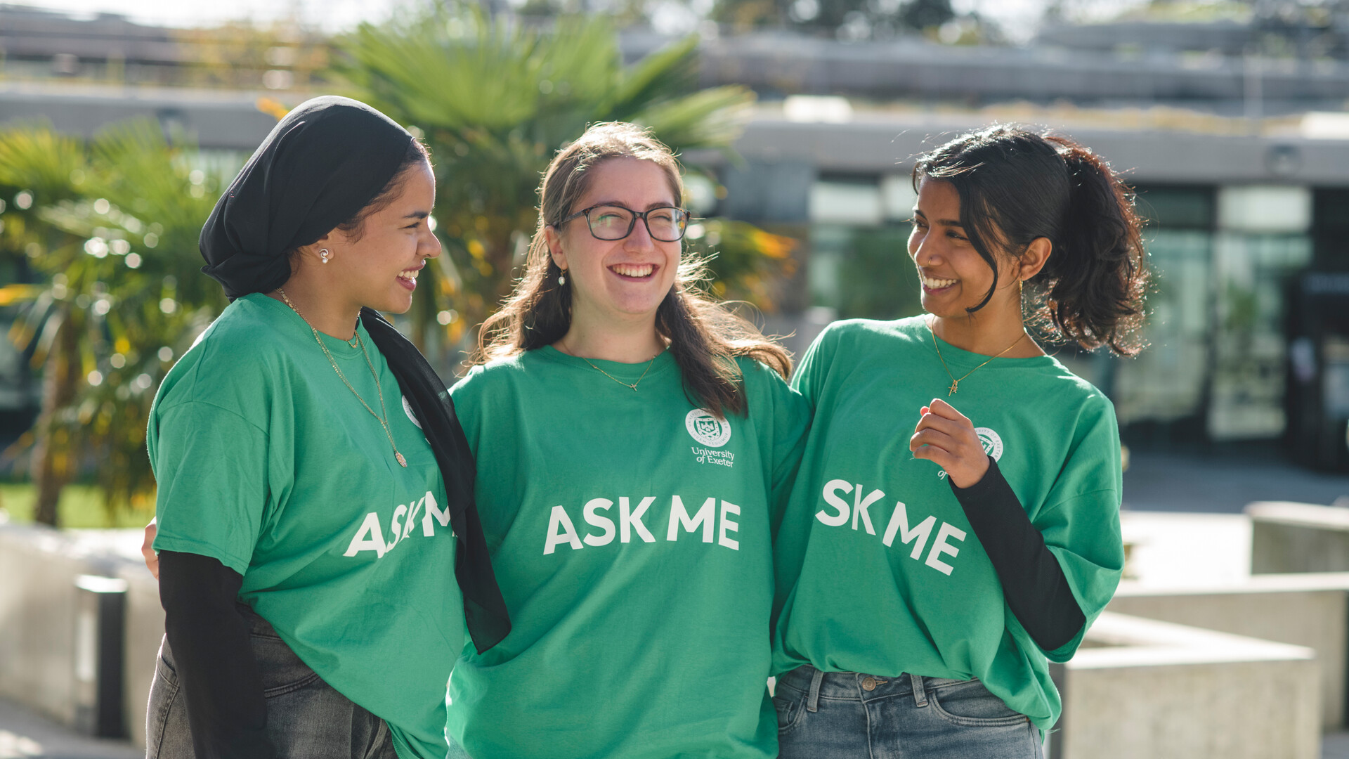 Three students in green university of Exeter tshirts that have 'ask me' in white writing. They are outside smiling at each other during a campus open day