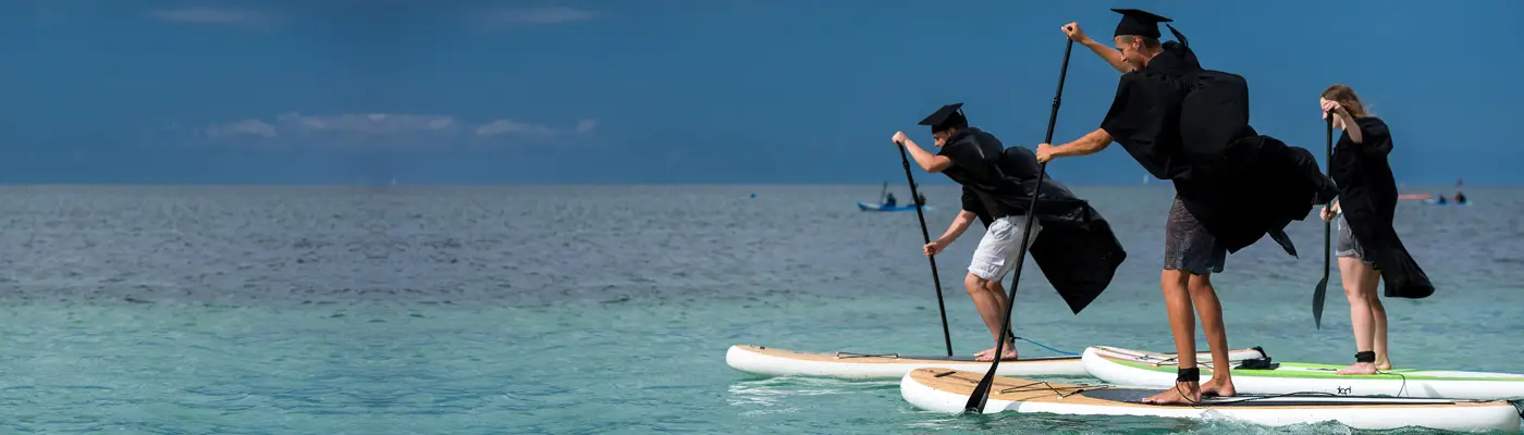 Two students on paddleboards in the sea wearing graduation gowns and mortarboards.