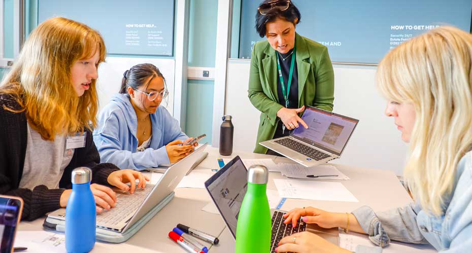 Students sitting at a table looking at a laptop that a lecturer is holding.