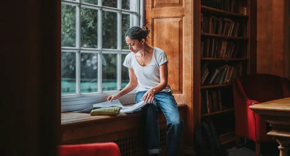 A female students sitting in a window seat looking at a book in a room full of bookshelves.