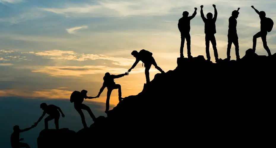 Silhouetted people helping each other to climb a rock