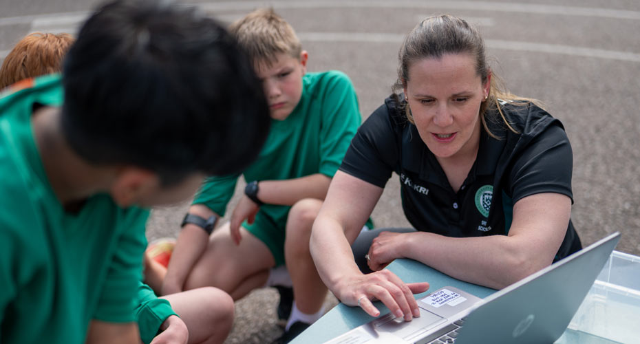 A researcher with school pupils gather round a laptop in a school playground