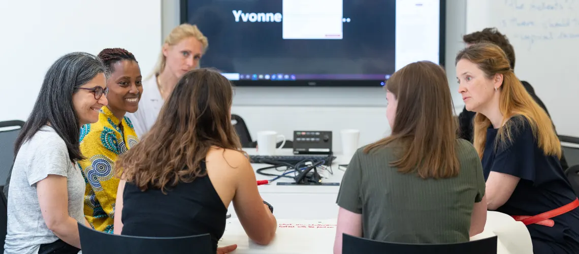 Group sitting around a white desk having a discussion, with a large monitor in the background.
