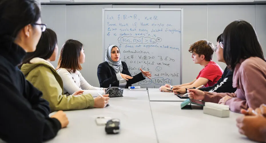 Maths lecturer teaching a group of students. There is a whiteboard in the background with mathematical equations on.