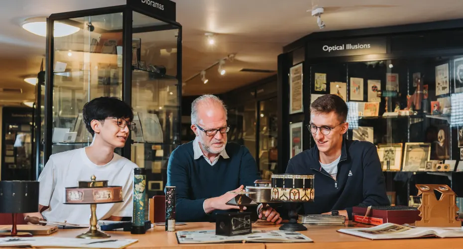 Two students and a lecturer in the Bill Douglas Cinema Museum, sitting on a table together examining museum artefacts.