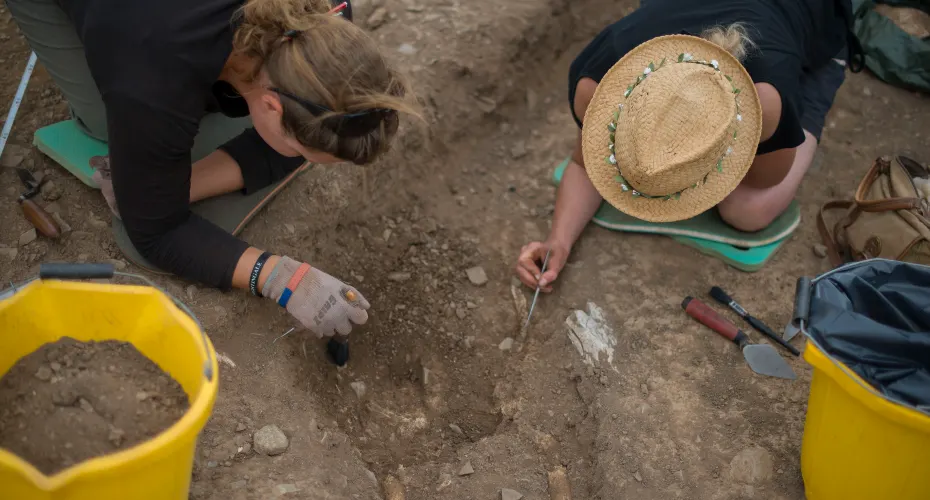 A view from above of two students kneeling on the ground, digging into soil during an archaeology fieldtrip.