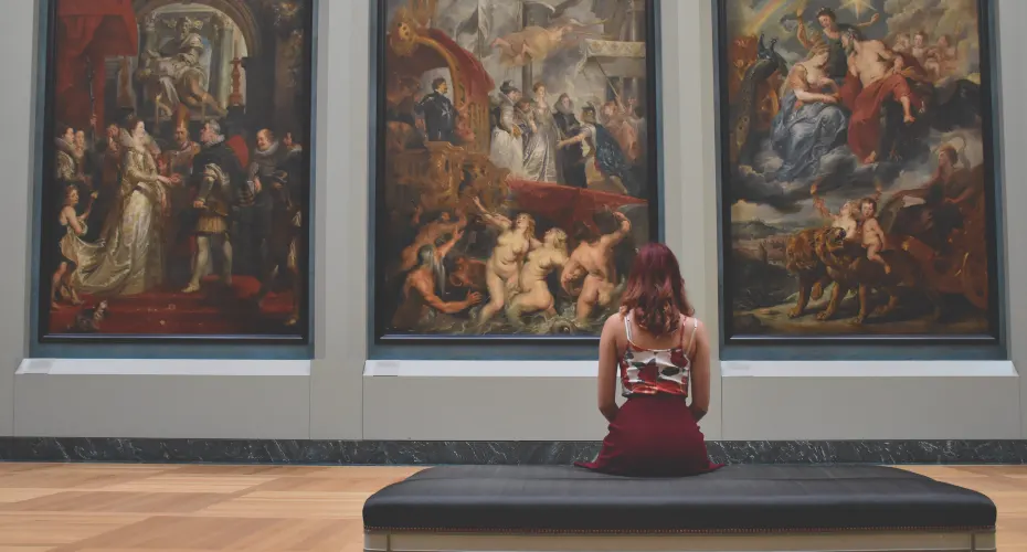 Person sitting in a gallery looking at three large historical paintings on the wall.