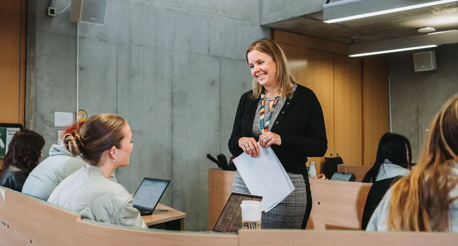 A lady who is smiling, standing at the front of a lecture theatre in front of students.