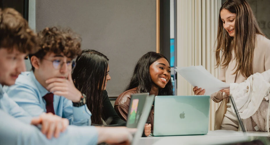 image of a group of students sat around a table. Three students can be seen smiling, while two other students look down at a laptop