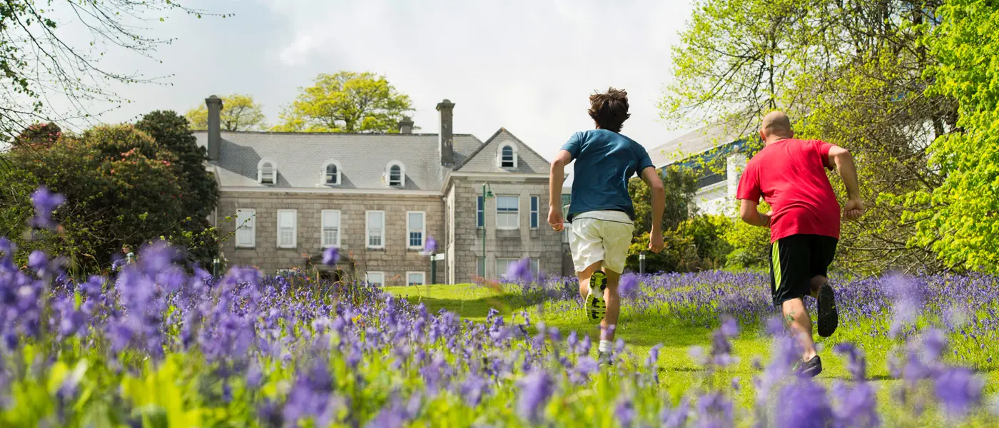 Two people running past the bluebells in front of Tremough House at our Penryn Campus.