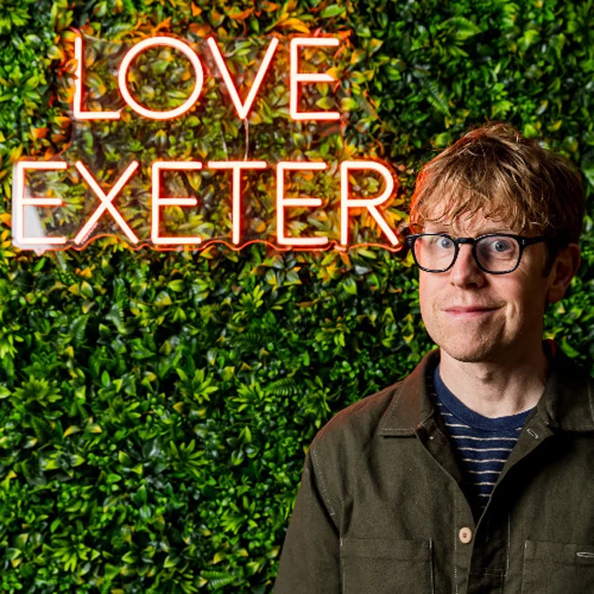 Josh Widdicombe standing in from our a green hedge with an iluminated sign that reads Love Exeter.