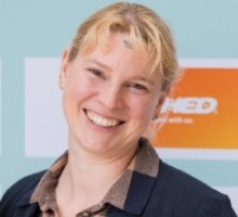 Judith has brown eyes and straight blonde hair. She wears a dark jacket, an orange and blue shirt and multiple stud earrings