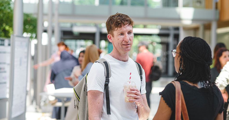 A man with curly hair and a backpack holds a drink with a straw, talking to a woman in a busy hallway.