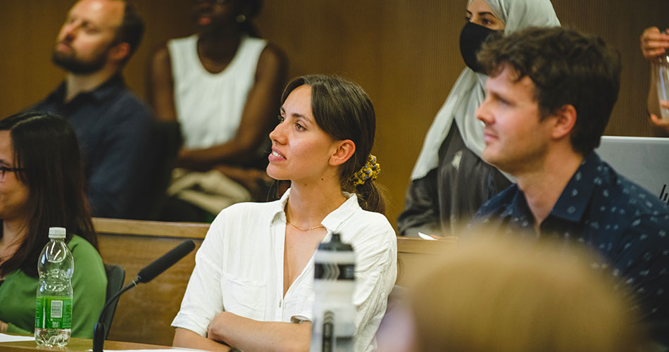 A woman with her hair tied back, wearing a white shirt, sits attentively in a room with other people, possibly during a meeting or lecture.
