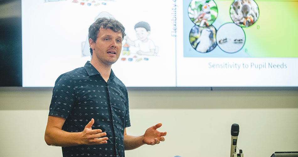 A man in a patterned short-sleeve shirt stands and presents in front of a screen displaying a slide titled 