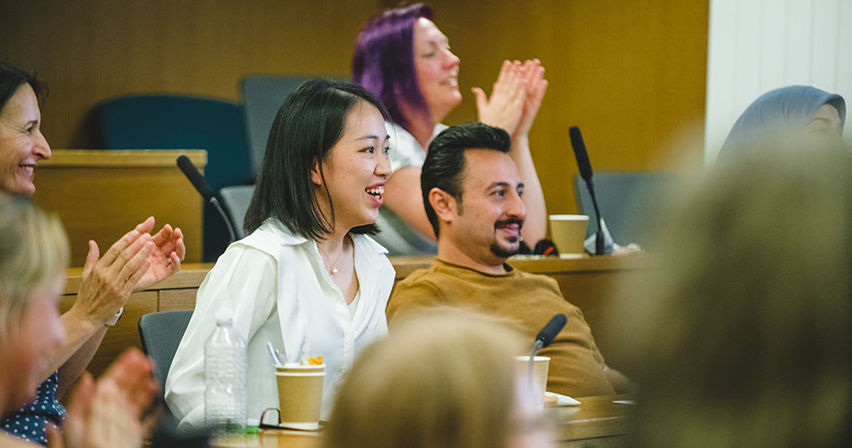 A group of people sitting in a lecture hall or conference room, clapping and smiling, with one woman in a white blouse prominently visible in the foreground.