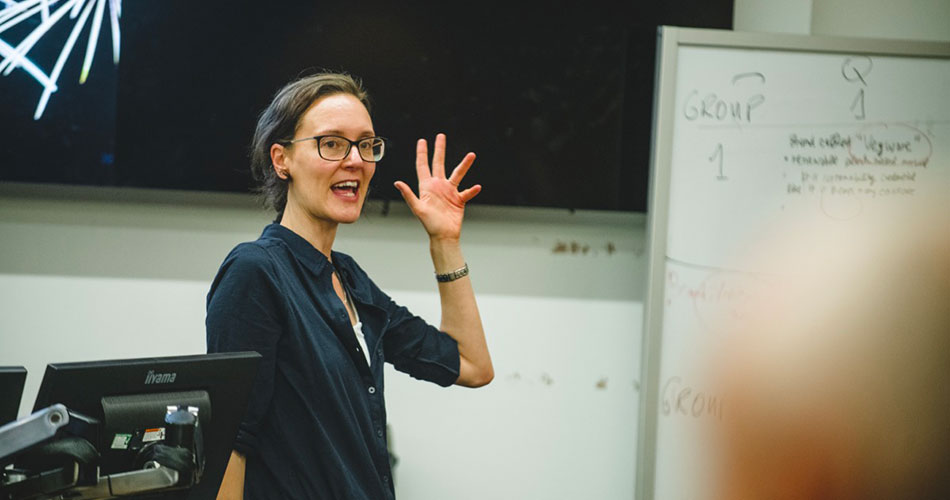 A woman with glasses and a dark blue shirt stands in front of a whiteboard, waving and smiling during a presentation.