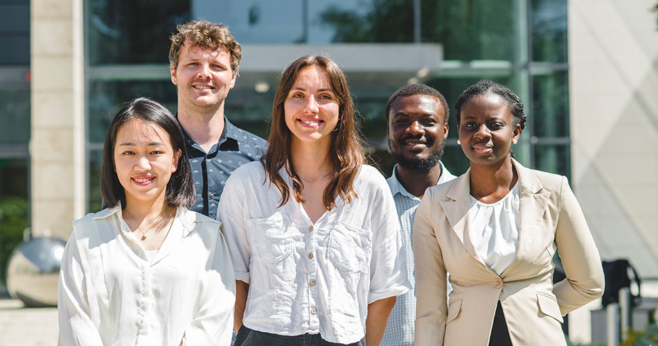 A group of five people standing outdoors in front of a modern building, smiling and posing for a photo.