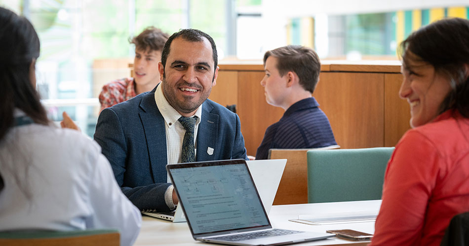A man in a suit and tie smiles while sitting at a table with a laptop, engaged in a discussion with three other people in a bright, modern indoor setting.