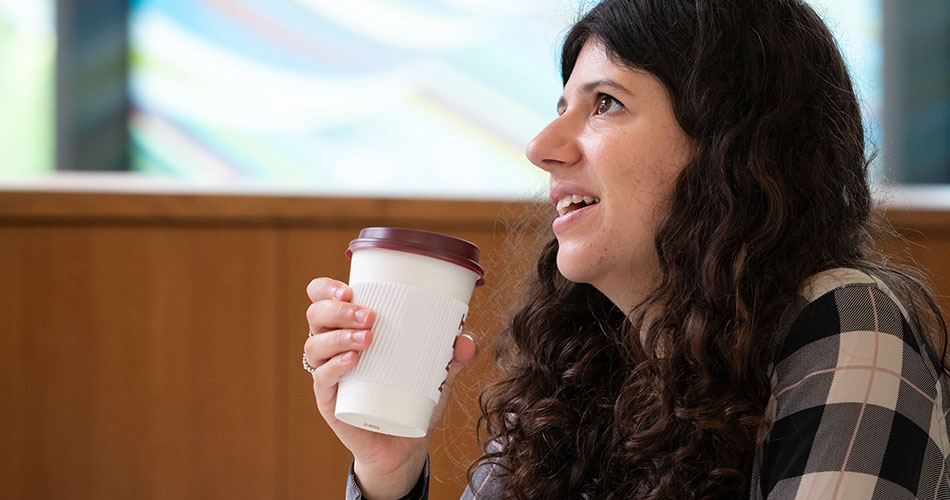 A woman with curly hair holding a coffee cup, smiling while looking off to the side.