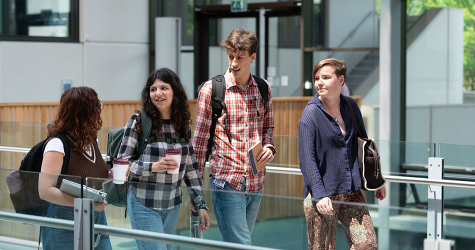 Four people walking outdoors on a campus, two holding coffee cups, engaged in conversation.