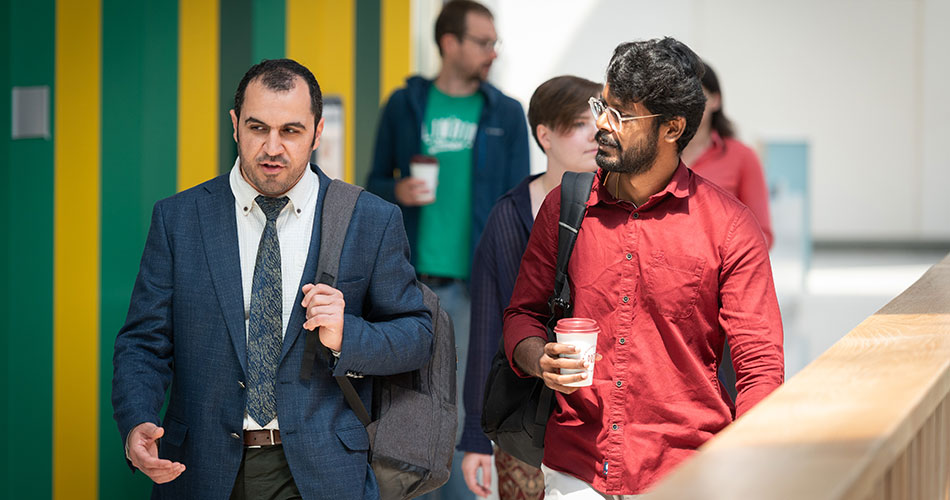 Two men walking indoors, one in a suit and tie holding a bag, the other in a red shirt holding a coffee cup.