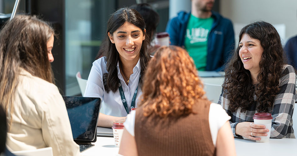 A woman with a lanyard smiles while engaging in a conversation with three other people seated around a table with laptops and coffee cups.
