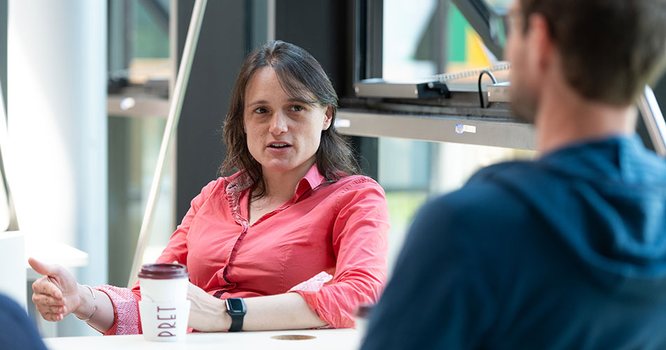A woman in a pink shirt holding a coffee cup, engaged in conversation with a man in a blue hoodie, both seated at a table indoors.