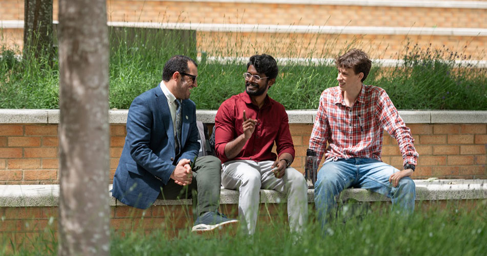 Three men sitting outdoors on a low wall, smiling and talking, surrounded by greenery.