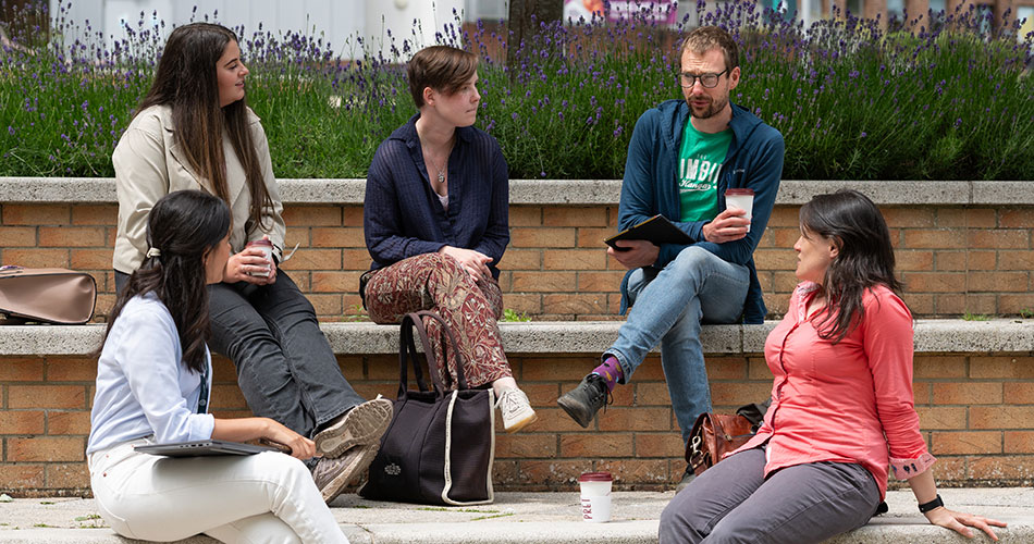Four people sitting outdoors on a low wall, engaged in conversation with coffee cups and notebooks, surrounded by lavender plants.