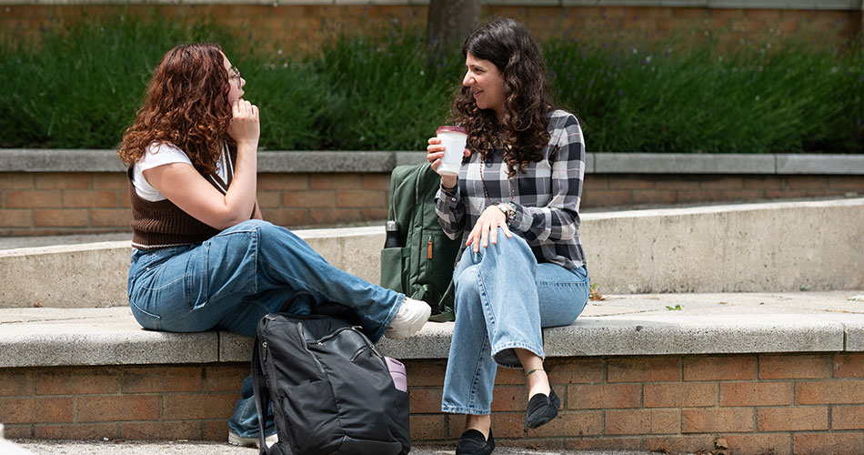 Two women sitting on a concrete ledge outdoors, one holding a coffee cup, engaged in conversation with a backpack and a bag beside them.