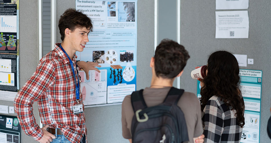 A man in a plaid shirt with a lanyard points to a poster during a presentation, explaining to two people.