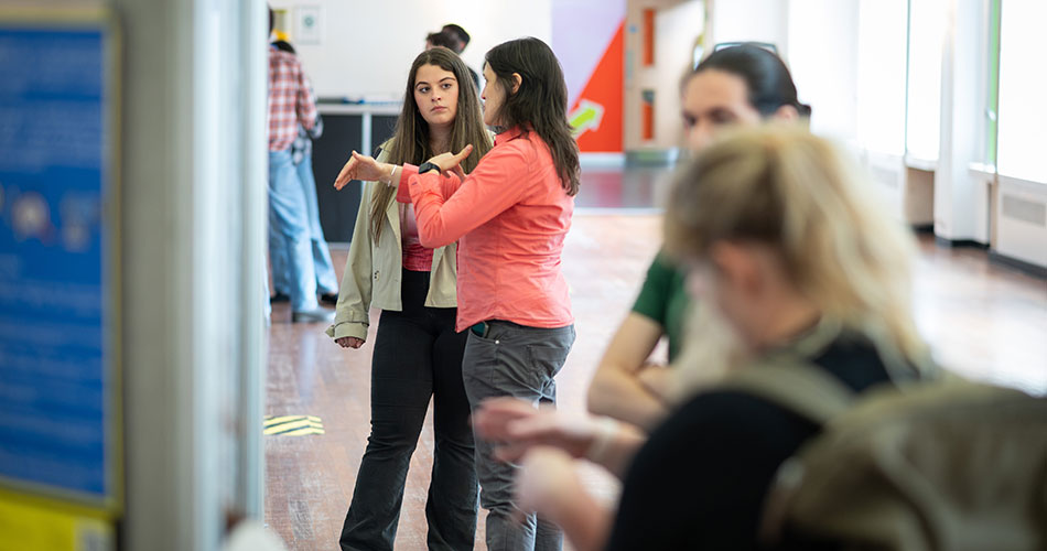Two women standing indoors, one gesturing with her arms crossed, engaged in conversation with others in the background.